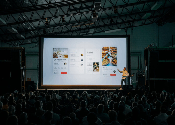 person discussing while standing in front of a large screen and audience inside dim-lighted room