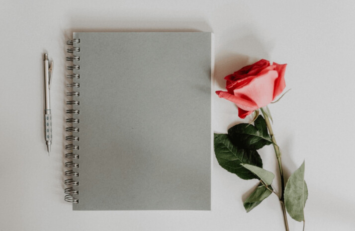 An orange rose flower beside a gray-colored notebook and pen