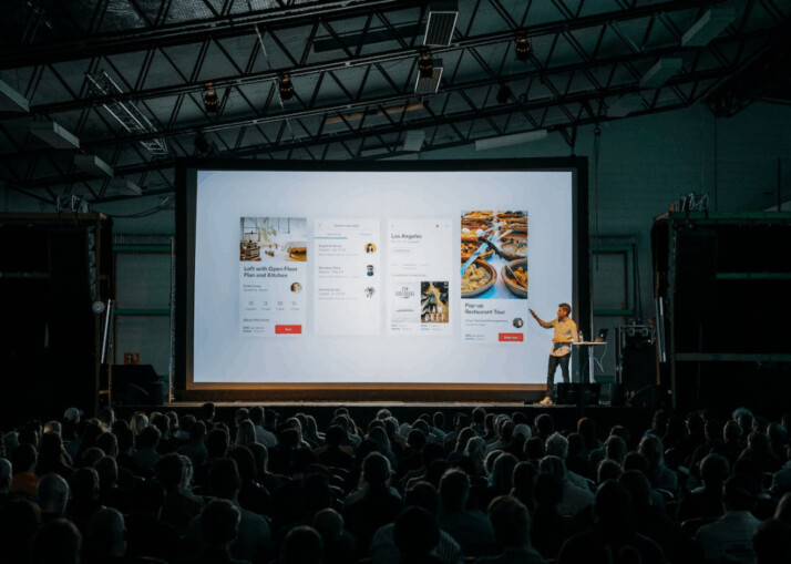 person discussing while standing in front of a large screen in front of people 