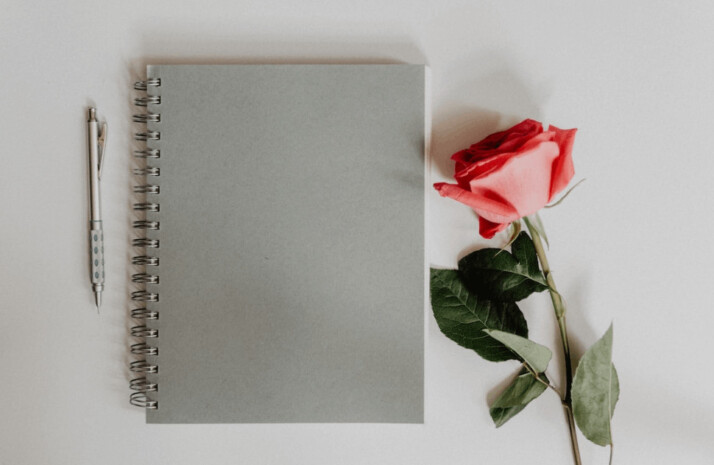An orange rose flower beside a gray notebook and pen
