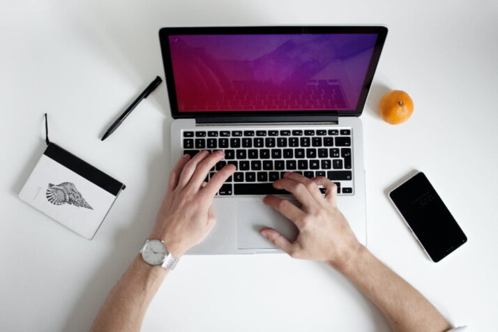 A person working on their laptop with a phone and orange next to it.