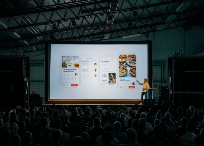 person discussing while standing in front of a large screen in front of people.