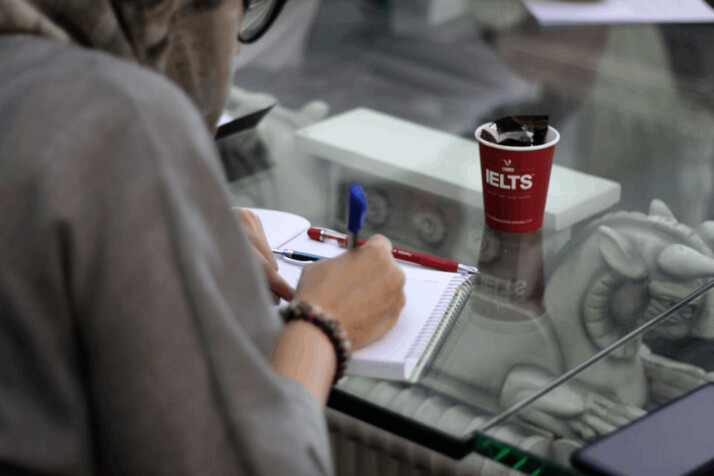 a person writing on paper and an IELTS paper cup placed on the table