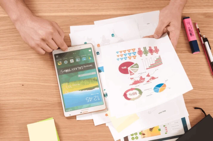 person holding a white Samsung Galaxy Tab with graphs and charts on paper.