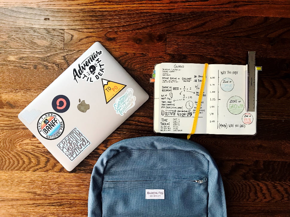 A backpack next to an open notebook and a laptop covered with stickers.