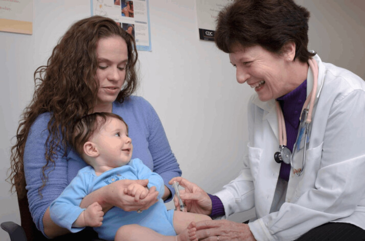 pediatrician smiling and pushing an injection on a little child smiling on his mother's lap