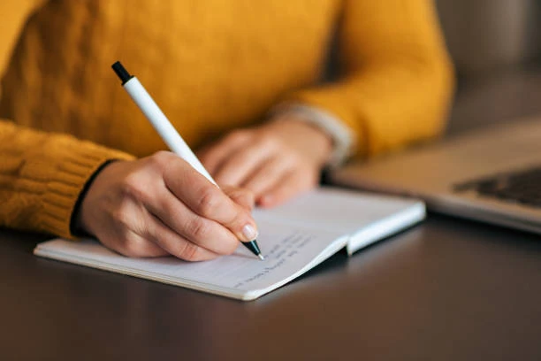 person wearing yellow sweater writing with white pen in notebook