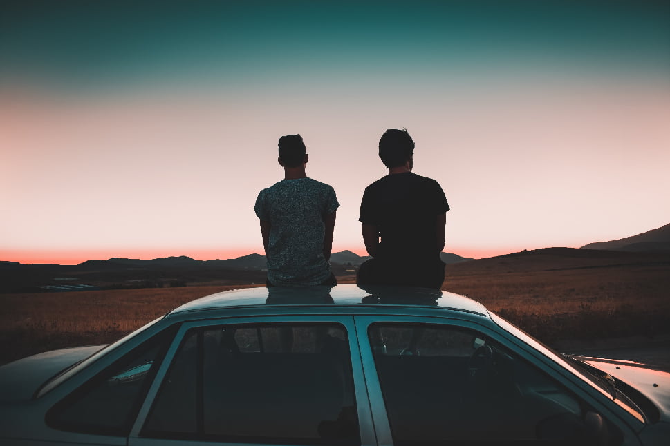Two guys sitting on the roof of a car while looking at the mountains.