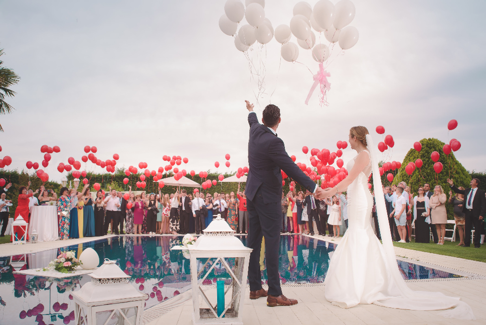 A wedding by the pool with the couple and guests holding out balloons.