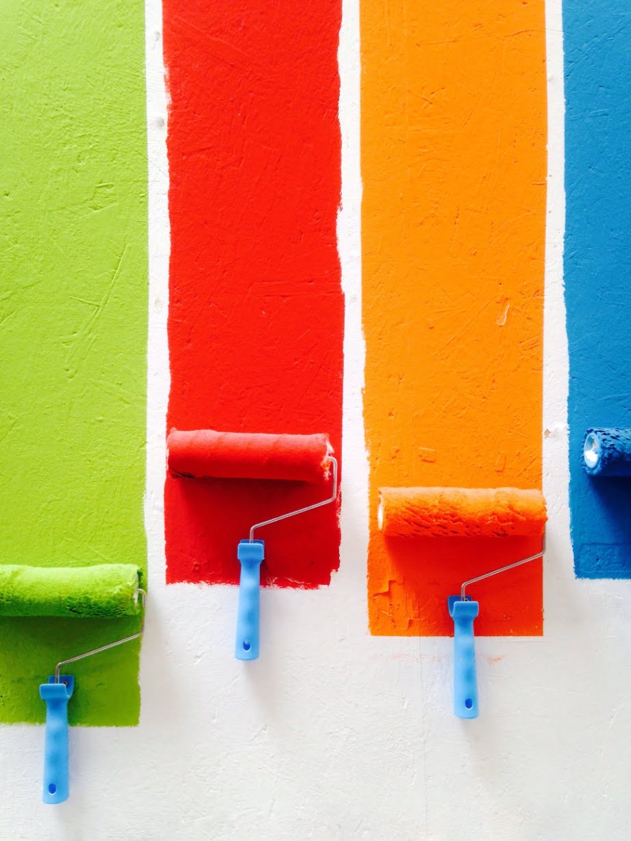 4 paint brushes, green, red, orange and blue painting a white wall.