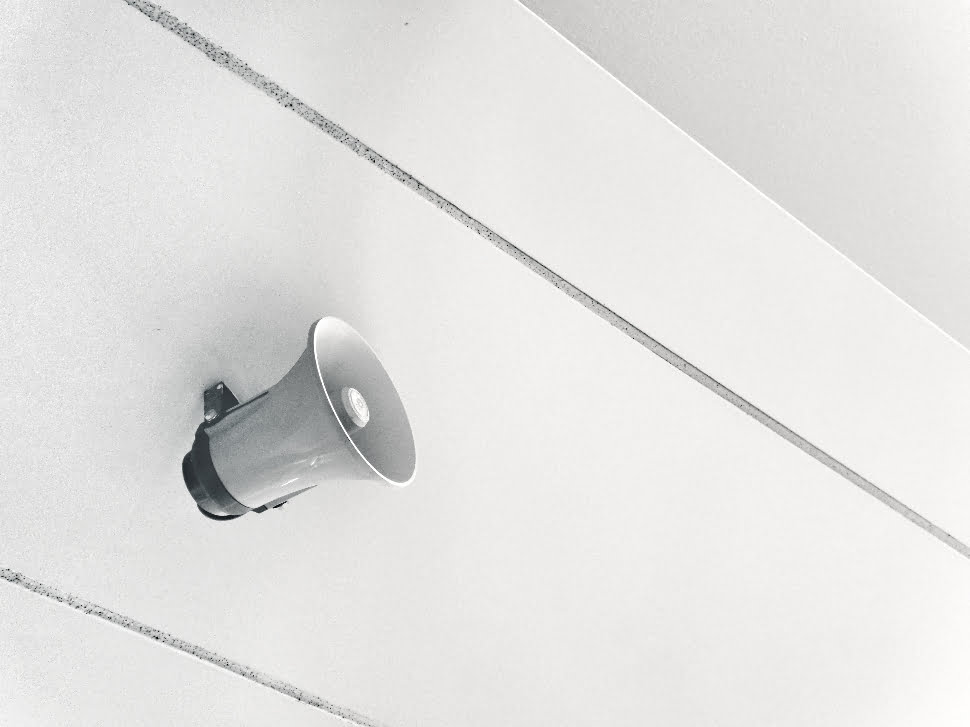 A grey speaker attached to the ceiling of a white building.