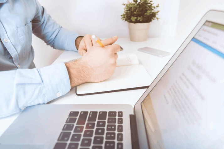 A person writing on a white notebook with a laptop opened on the side