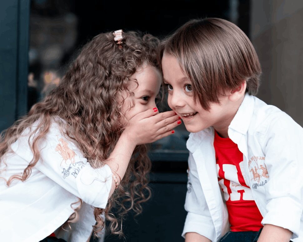 A little girl whispering in a little boy's ear. 