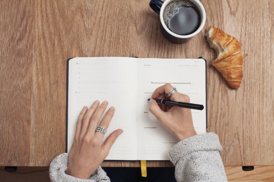 A person writing on a notebook with a croissant and cup of coffee beside them.