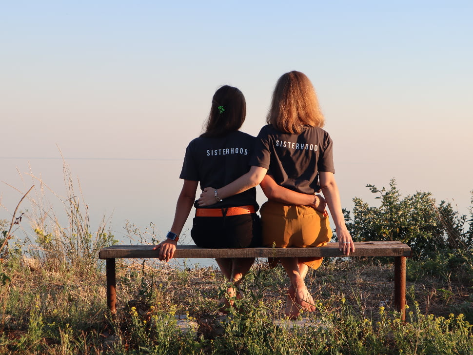two women hugging each other facing the sea with tshirts that read 