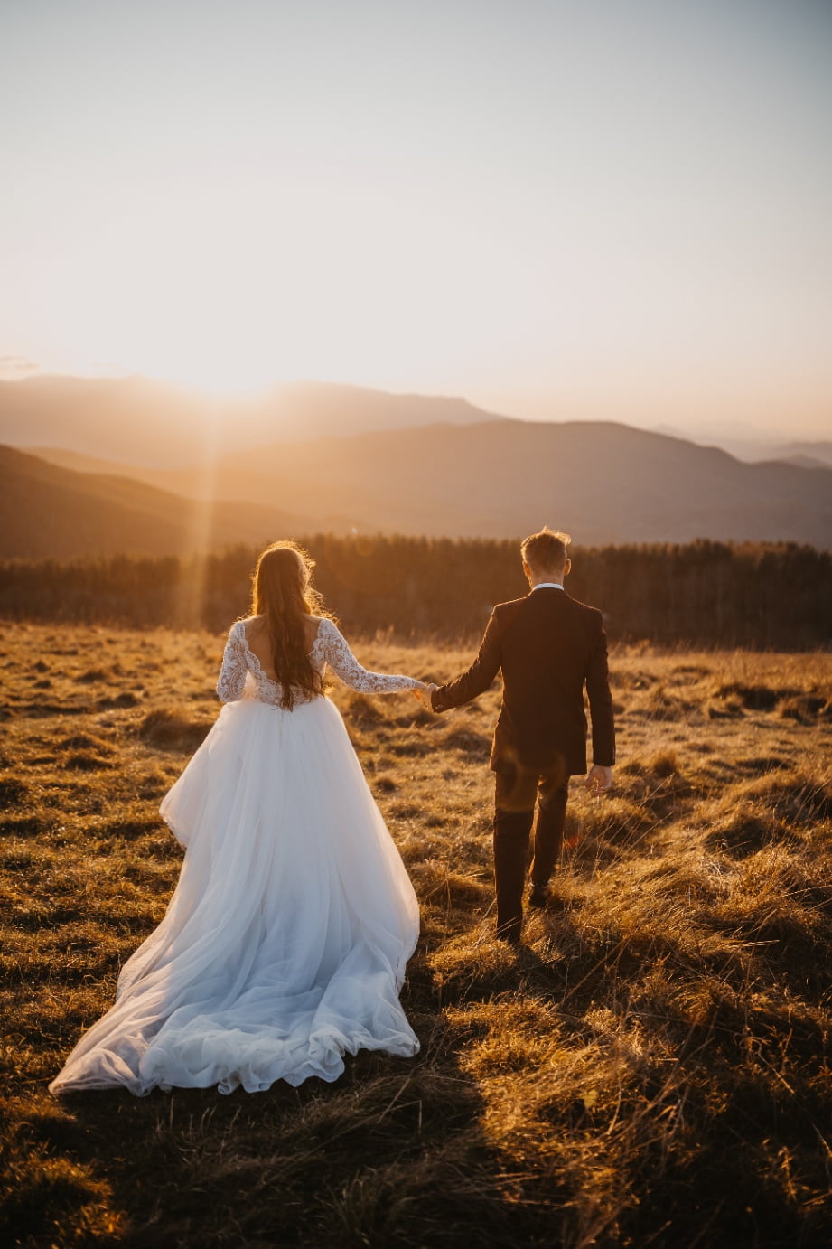 A bride and groom walking through grass towards the sunset.