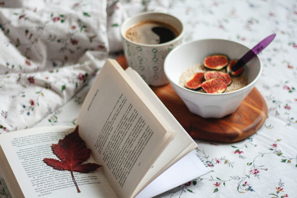 An open book with a leaf bookmark next to a bowl of food and coffee.