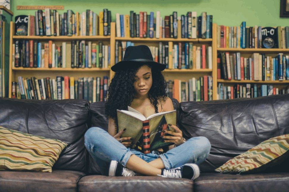 Curly-haired woman engrossed in a book while seated on a sofa, bookshelf adding to the ambiance