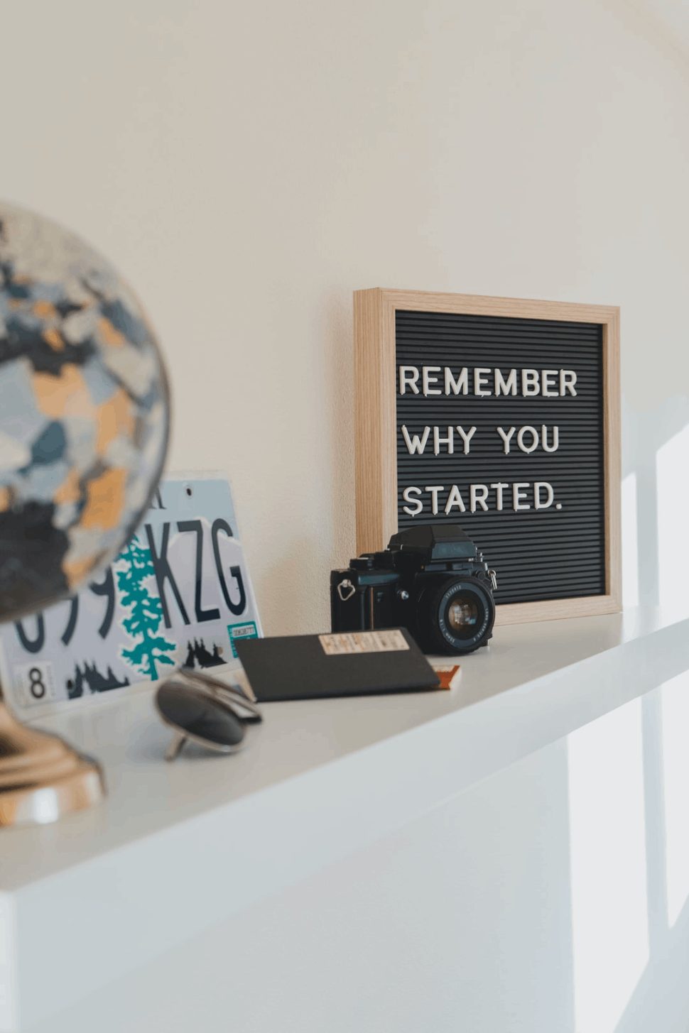Motivational reminder, "Remember why you started," placed on a table with camera, passport, and glasses