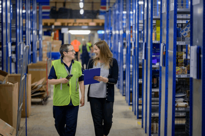 Two women in a warehouse walking down a path talking to each other and smiling.