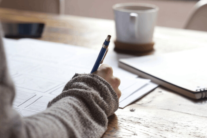 Person writing on brown wooden table near white ceramic mug