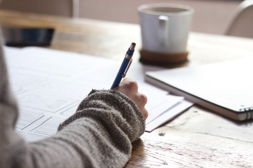 A hand holding pen, writing on paper with a coffee mug in the background