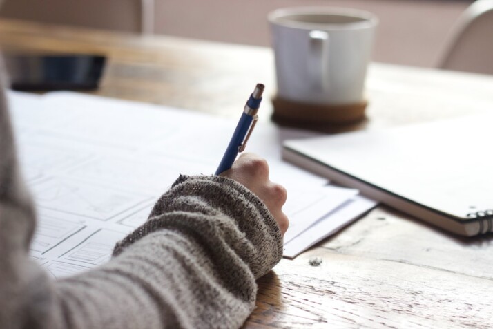 A person writing on brown wooden table near a white ceramic mug.